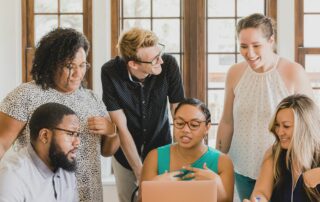 six men and women of different ethnicities sitting around a laptop discussing what is on the screen