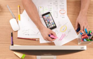 downward view of desk covered in papers with pens, highlighters, and a cell phone spread out in front of a desktop computer
