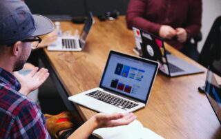 Downward view of four people at a table with laptop open. Man in focus with plaid shirt and baseball cap has his hands upraised in a shrug